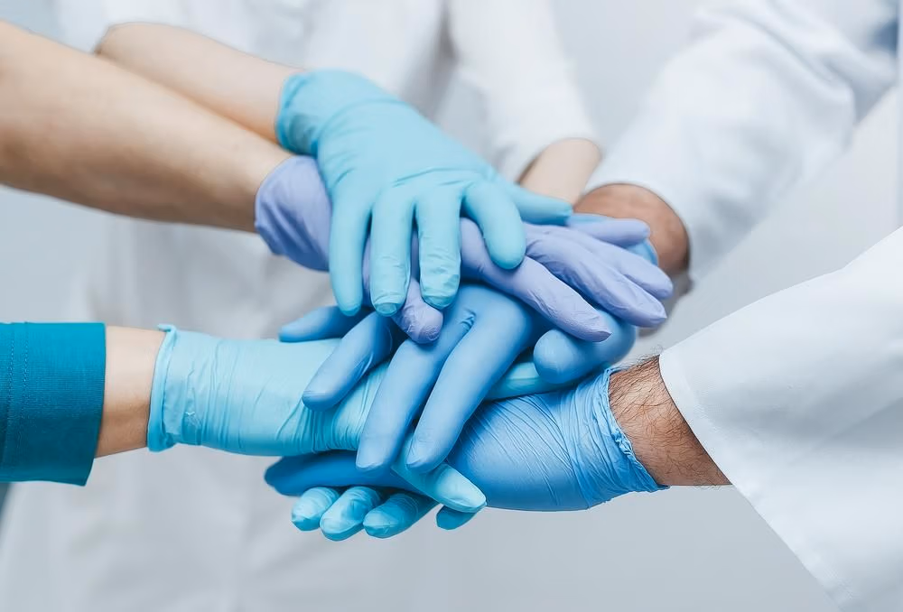 Team,Of,Young,Doctors,Stacking,Hands,In,Blue,Gloves,Indoor. Team,Of,Young,Doctors,Stacking,Hands,In,Blue,Gloves,Indoor.